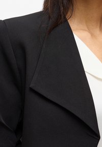 Svart blazer med en strukturerad slag och något texturerat tyg, delvis synlig tillsammans med en vit topp.