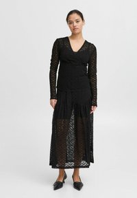 Abito lungo in pizzo nero con maniche lunghe e scollatura a V, caratterizzato da una gonna a strati e tessuto trasparente. Indossato con ballerine nere a punta.