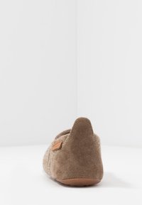 Chausson en daim marron avec une surface texturée, un bout arrondi et un petit talon. Présente une étiquette d'accent beige près du talon.