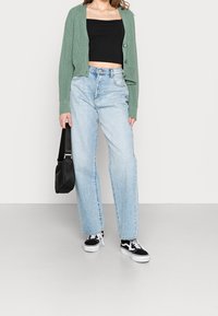 Žena v zeleném pleteném kardiganu, černém crop topu, světle modrých vysokých džínách, černo-bílých teniskách a s černou taškou.