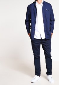 Mörkblå skjorta med knappar och ficka, lagd över en vit skjorta, matchad med mörkblå byxor och vita sneakers. Slät struktur, skräddarsydd passform.