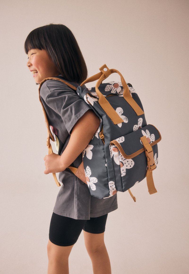 Grauer Stoffrucksack mit Blumenmustern, braunen Riemen und einer Vordertasche. Der Rucksack verfügt über einen oberen Griff und gepolsterte Riemen für Komfort.
