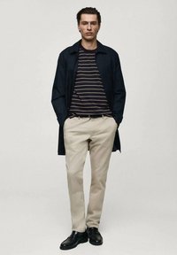 Marinblå långkappa över randig svartvit t-shirt, beige byxor och svarta skor. Personen står med händerna i fickorna, neutral bakgrund.