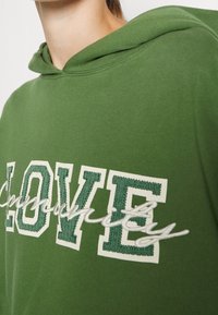 Groene katoenen hoodie met geribbelde manchetten, een voorzak en het woord "LIEFDE" in vetgedrukte witte en groene borduursel.