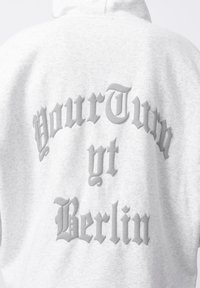 Siva kapuca z zmehko teksturo in dvignjenim napisom na hrbtni strani, ki pravi "YourTurn yt Berlin" v kontrastni sivi pisavi.