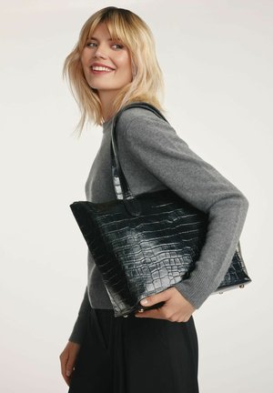 Blondynka uśmiechająca się, ubrana w szary sweter i czarne spodnie, nosząca dużą czarną torebkę o fakturze krokodyla na ramieniu na białym tle.