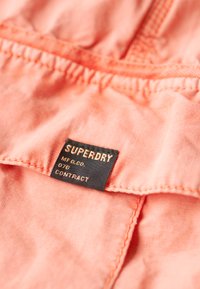 Koraal stof met een gestructureerde afwerking, featuring een zwart label met gouden tekst met de opschrift "SUPERDRY MFG. CO. 076 CONTRACT." Stiksels zichtbaar.
