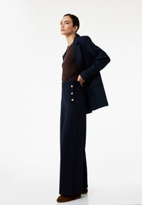 Navy wijde broek met gouden knoopaccenten op de zakken, gecombineerd met een bruine top en een marineblauwe blazer, ontworpen met een getailleerde pasvorm.