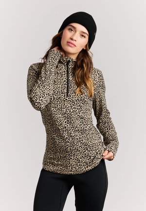 Vrouw die een lange mouwen top met luipaardprint en duimgaten draagt en een zwarte muts, poserend tegen een egale lichte achtergrond.