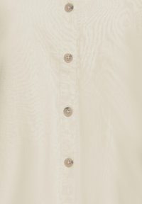 Közeli kép egy bézs színű anyagpanelről, amelyen egy függőleges sorban négy áttetsző, bézs színű gomb található, mindegyiken négy lyukkal varrva.