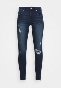 Skinny jeans in dark blue denim with distressed accents. Features a mid-rise waist, five pockets, and ankle zippers for added detail.