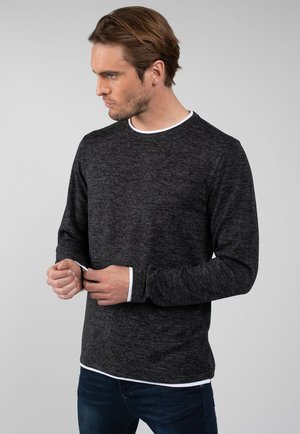 Mand med kort brunt hår iført en mørkegrå langærmet skjorte over en hvid undertrøje, justerer sin manchet, vender mod venstre.