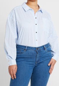 Camisa de botones a rayas en azul claro y blanco, con un bolsillo en el pecho y mangas largas. Detalles de botones en los puños. Combinada con jeans azules.