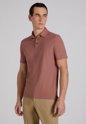 Uomo che indossa una polo a maniche corte color rosa polvere e pantaloni beige, guarda verso sinistra su uno sfondo grigio uniforme.