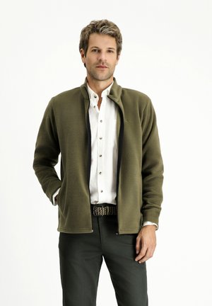 Giacca in pile verde oliva con colletto alto, zip nera e cuciture decorative. Indossata sopra una camicia bianca con bottoni e pantaloni scuri.