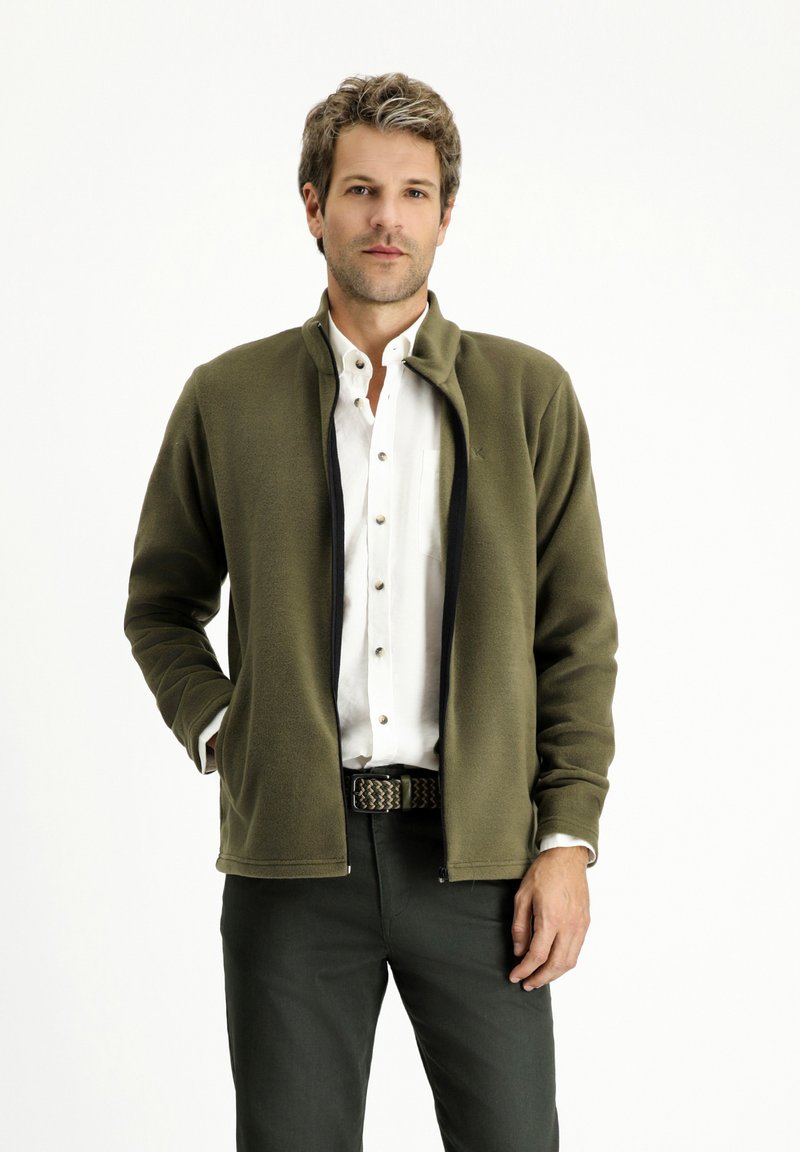 Giacca in pile verde oliva con colletto alto, zip nera e cuciture decorative. Indossata sopra una camicia bianca con bottoni e pantaloni scuri.