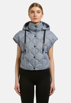 Junge Frau trägt eine ärmellose hellblaue gesteppte Kapuzenweste mit schwarzen Kordeln und schwarze Hose.