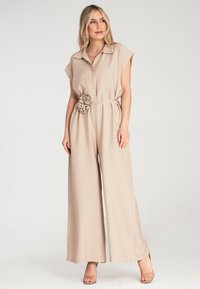 Beżowy kombinezon z krótkimi rękawami, kołnierzykiem, guzikami z przodu, cienkim talią i materiałowym detalem w postaci kwiatu w talii. Szerokie nogawki.