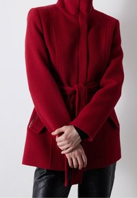 Abrigo de mezcla de lana roja con un alto cuello, cintura ceñida por un cinturón y bolsillos laterales con detalles de cremallera; forma estructurada y textura suave.