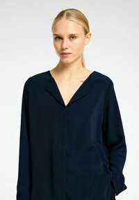 Marineblå langærmet bluse med V-hals og afslappet pasform. Fremstillet af glat stof med minimal detaljer og ingen synlige lukninger.