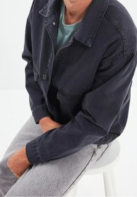 Tmavě modrá džínová bunda s knoflíkovým zapínáním a dvěma kapsami na prsou, nošená přes světle zbarvené tričko. Šedé kalhoty, sedící na bílém stoličce.