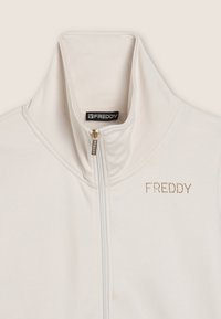 Giacca con zip color crema e collo alto, etichetta del marchio "FREDDY" all'interno del collo, e testo "FREDDY" in piccole pietre preziose sul petto sinistro.