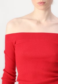 Czerwona bluzka typu off-shoulder z dzianiny o prążkowanej strukturze i długimi rękawami; posiada dopasowany krój oraz fakturowany materiał. Bez widocznych wzorów ani elementów metalowych.