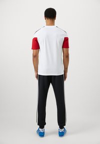 Herre atletisk outfit bestående af en rød kortærmet top, hvid krop og sorte bukser med hvide striber. Skoene er blå. Materialet er en blanding af bomuld.