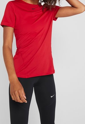 Vrouw die een rood Nike T-shirt met korte mouwen en zwarte Nike legging draagt, poseert met één arm omhoog tegen een effen achtergrond.