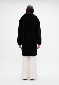 Palton oversized, negru și pufoș, cu guler, mâneci lungi și tiv drept, purtat cu pantaloni albi largi. Vedere din spate fără alte detalii.