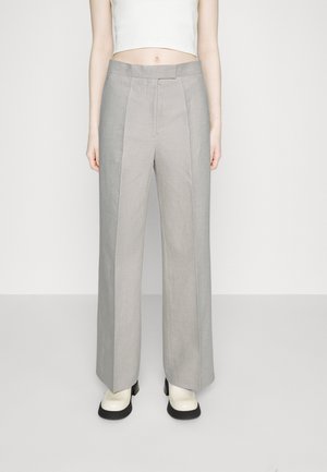 Vrouw draagt lichtgrijze, hooggetailleerde, wijde broekspijpen, wit cropped top en witte plateauzolen met zwarte zolen tegen een effen achtergrond.