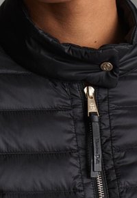 Veste matelassée noire avec un col montant, des détails en métal doré et un tirette de fermeture éclair contrastante. Texture lisse avec des motifs de couture horizontaux.