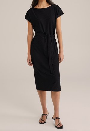 Schwarzes knielanges Kleid mit strukturiertem Muster, kurzen Ärmeln und gebundenem Taillengürtel. Kombiniert mit flachen schwarzen Sandalen.