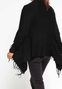 Svart poncho-stil cardigan med lös passform, långa ärmar, fransdetaljer längs kanten och en mjuk stickad textur.