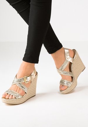 Sandales à talons compensés en métal doré avec des brides tissées et un détail de boucle, dotées d'une semelle plateforme en jute texturé, portées avec un pantalon noir ajusté.