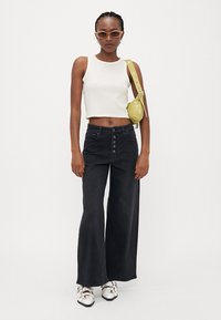 Čierne džínsy s vysokým pásom a širokými nohavicami s prednými gombíkmi, spárované s bielym crop topom a malou zelenou crossbody taškou. Strieborné tenisky dotvárajú vzhľad.