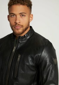 Sort læderjakke med opretstående krave, lynlås fortil og to sidelommer. Har en glat tekstur og subtil branding på ærmet.