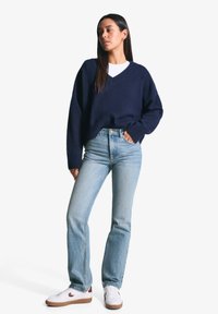 Mornarsko pleteno pulover s V-izrezom, kombiniran s svetlo modrimi jeans hlačami s ravnim krojem in belimi teniskami z rjavimi detajli.