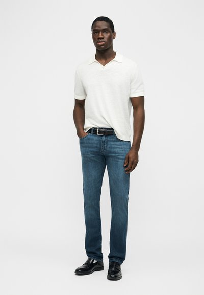 Νέος άνδρας που στέκεται με τα χέρια στις τσέπες, φορώντας λευκό polo με κοντά μανίκια, μπλε τζιν, μαύρη ζώνη και μαύρα παπούτσια σε απλό φόντο.