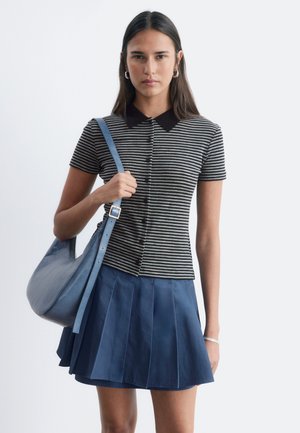 Jeune femme portant une chemise rayée noire et grise boutonnée, une jupe plissée bleu marine, et portant un sac à bandoulière bleu.