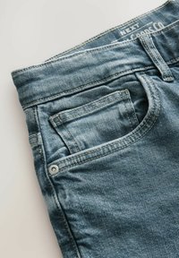 Jeanși din denim albastru cu o textură decolorată, având un buzunar frontal, talie curbată și cusături contrastante. Detaliu metalic pentru nasture vizibil.