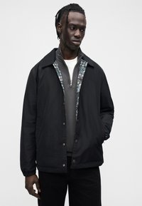 Casaco leve preto com fecho frontal de pressionar, forro estampado e corte descontraído. Combinado com um casaco cinzento com fecho de correr e calças pretas.