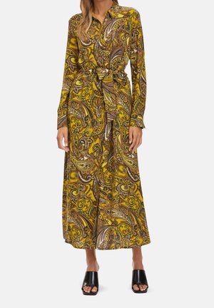 Vrouw draagt een gele midi-jurk met lange mouwen en paisleyprint, een taillestrik en zwarte open-teen blokhakmuiltjes.