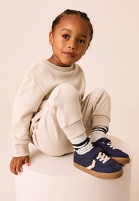 Sweat-shirt beige et joggings avec poignets côtelés. Baskets bleu foncé avec accents crème et semelles beige. Chaussettes à rayures noires et blanches.