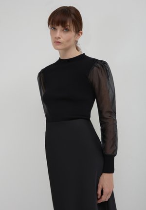 Top nero a maglia con maniche lunghe e trasparenti nere. Le maniche presentano un motivo sottile, e il top ha un design aderente con scollatura a girocollo.