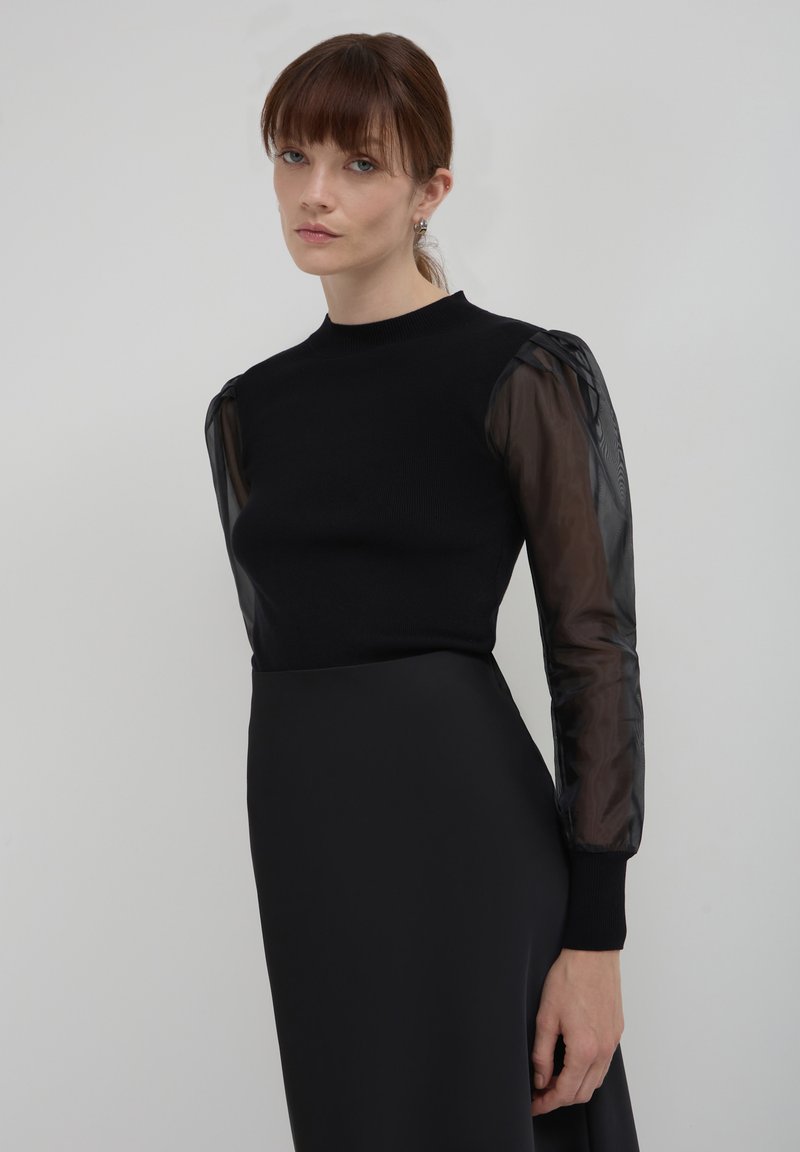 Top nero a maglia con maniche lunghe e trasparenti nere. Le maniche presentano un motivo sottile, e il top ha un design aderente con scollatura a girocollo.