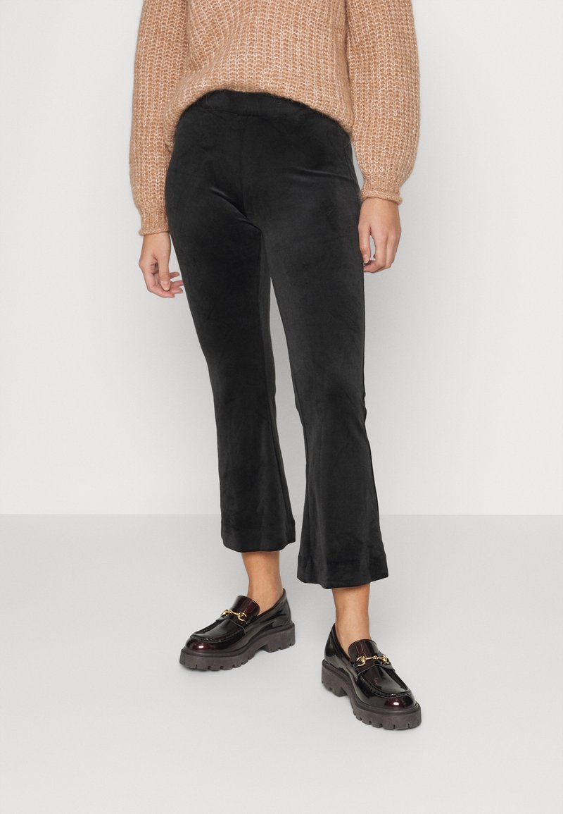 Femme portant un pantalon court en velours noir, un pull en maille couleur beige et des mocassins noirs épais avec un détail chaîne dorée sur un fond uni.
