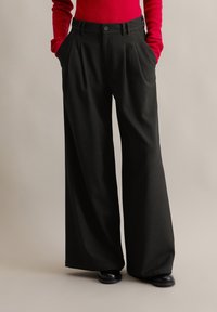 Pantalones de pierna ancha negros con pliegues, que presentan una textura suave y bolsillos, combinados con un suéter rojo ajustado.