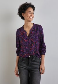 Blusa viola a fantasia con maniche lunghe, caratterizzata da un scollo a V e polsini arricciati, abbinata a jeans neri. Tessuto morbido con disegni intricati.