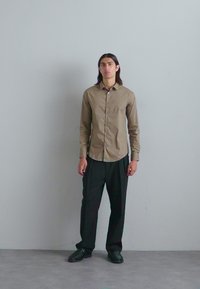 Chemise brune à boutons avec manches longues, portée avec un pantalon et des chaussures noires. Design simple, texture lisse, dotée d'une poche unique.